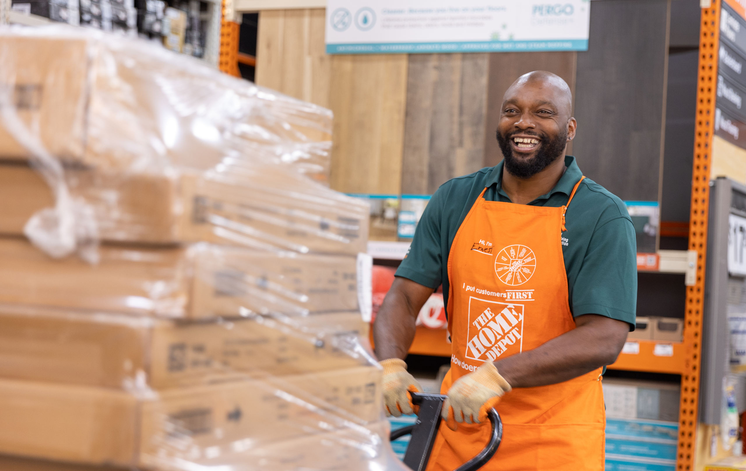 Middle-aged black man in a green polo and orange apron smiles as he transports boxes in a store.