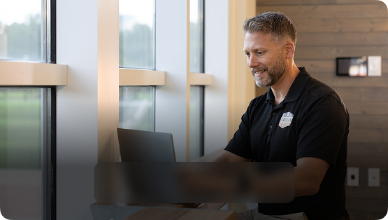 A man smiling while working on a laptop at a desk near a window