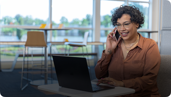 A woman smiling and talking on a cell phone while using her laptop in a modern office space